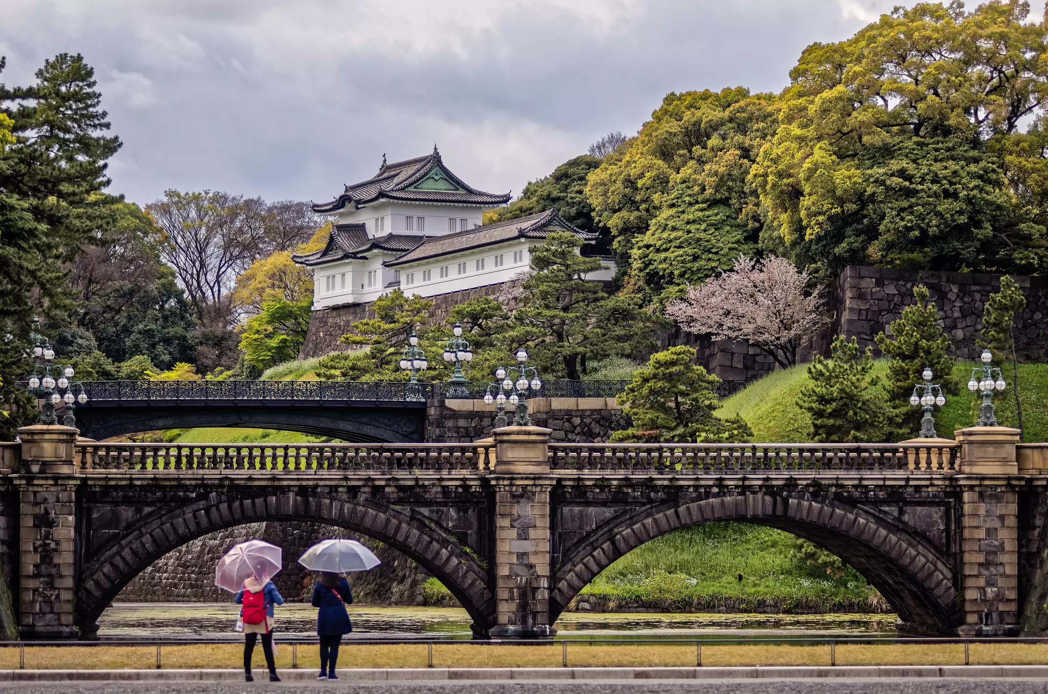 Two people holding umbrellas face a stone bridge and a Japanese palace on a hillside.