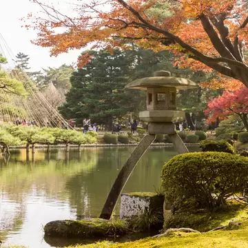 Two-legged stone lantern by a pond in Kenroku-en garden, Kanazawa, with rope-supported pines and an orange autumn maple