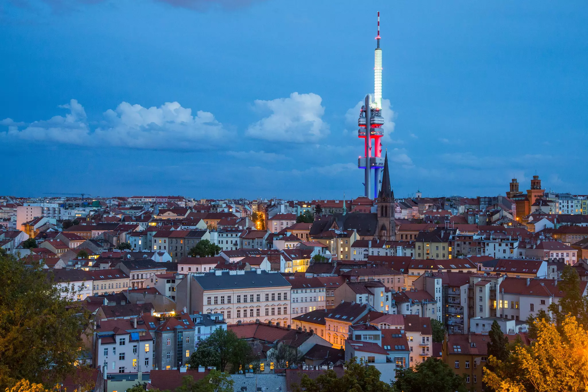 A large TV tower lit up in white, red and blue lights stands tall above residences in a low-rise city neighborhood.