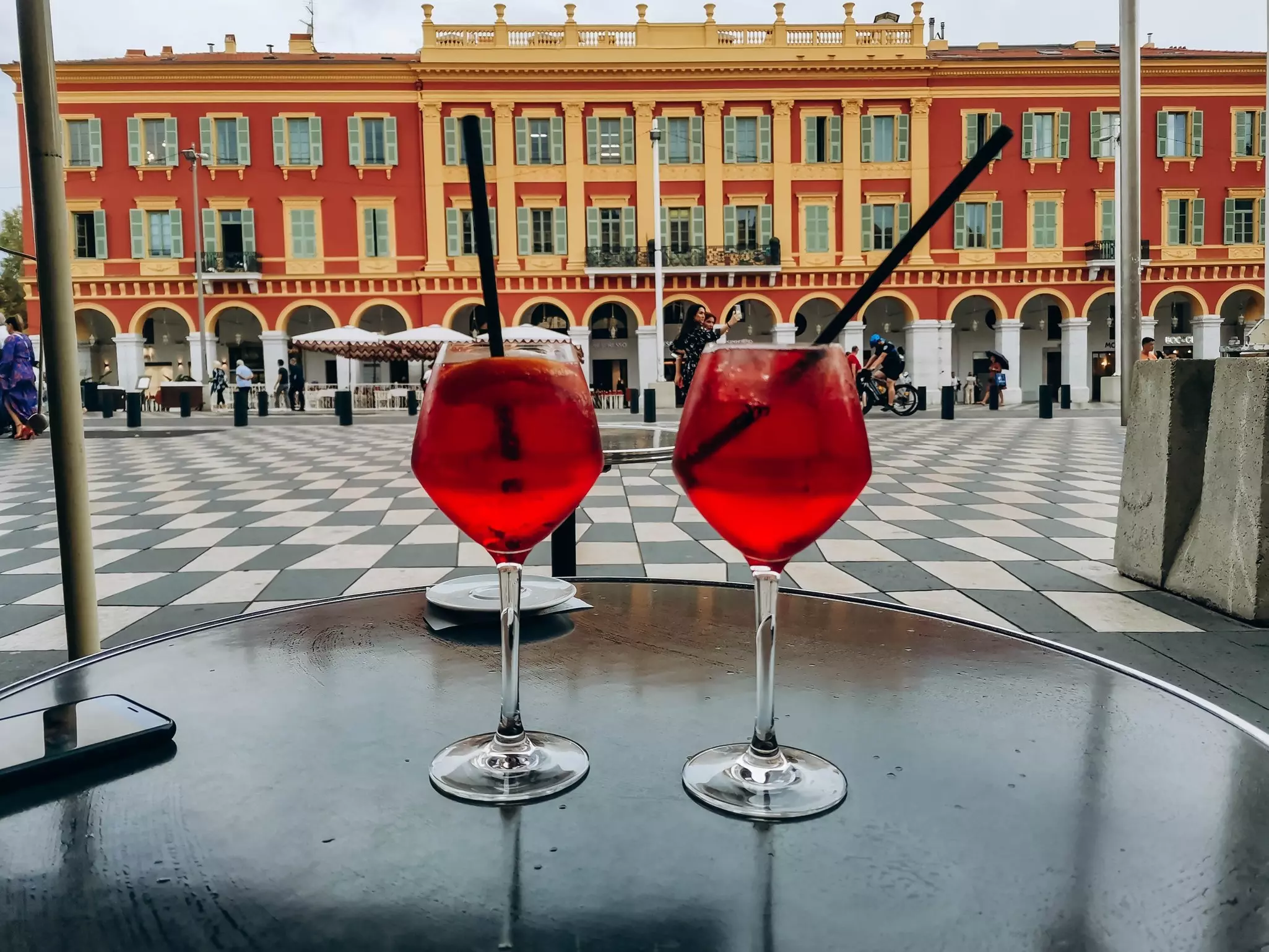 Two pink-colored cocktails sit on a table next to a city square with checkerboard pavers, and a colonnaded palace on the far side.