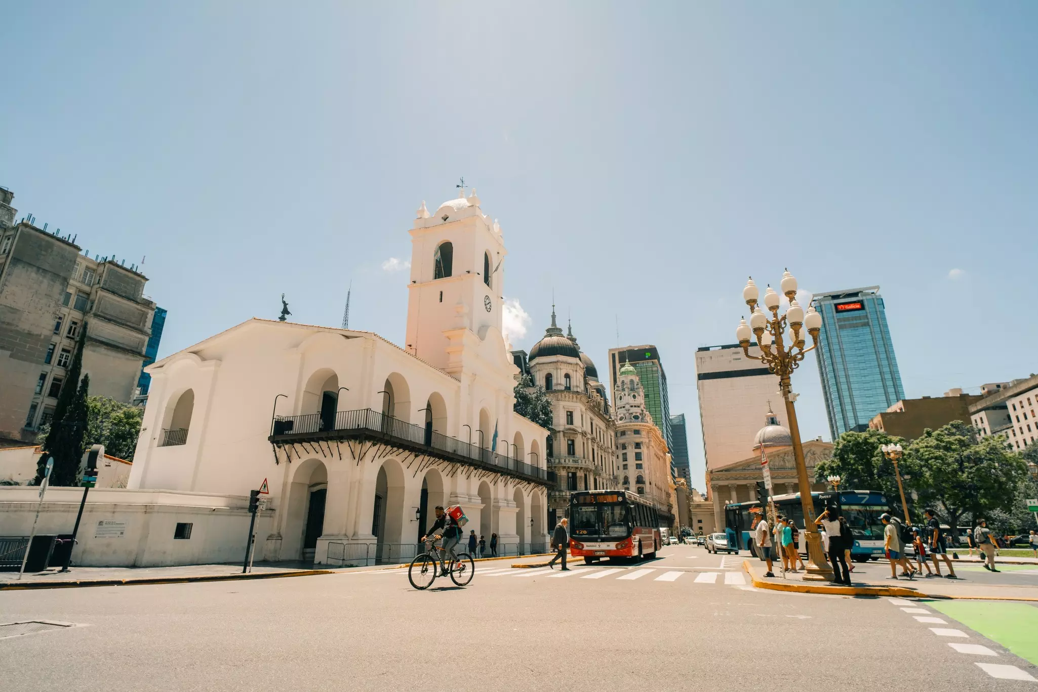 large white colonial building with clock in center tower and wrap around iron deck fronted by a street filled with people, a bus and bike.