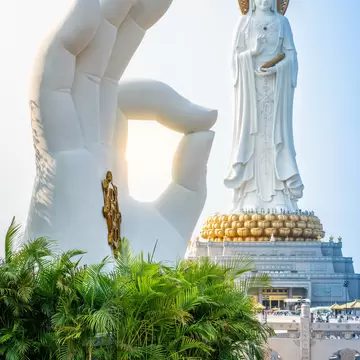 A giant white Guanyin statue on a golden lotus base beside a sculpted hand and palms at Nanshan, Sanya, Hainan.