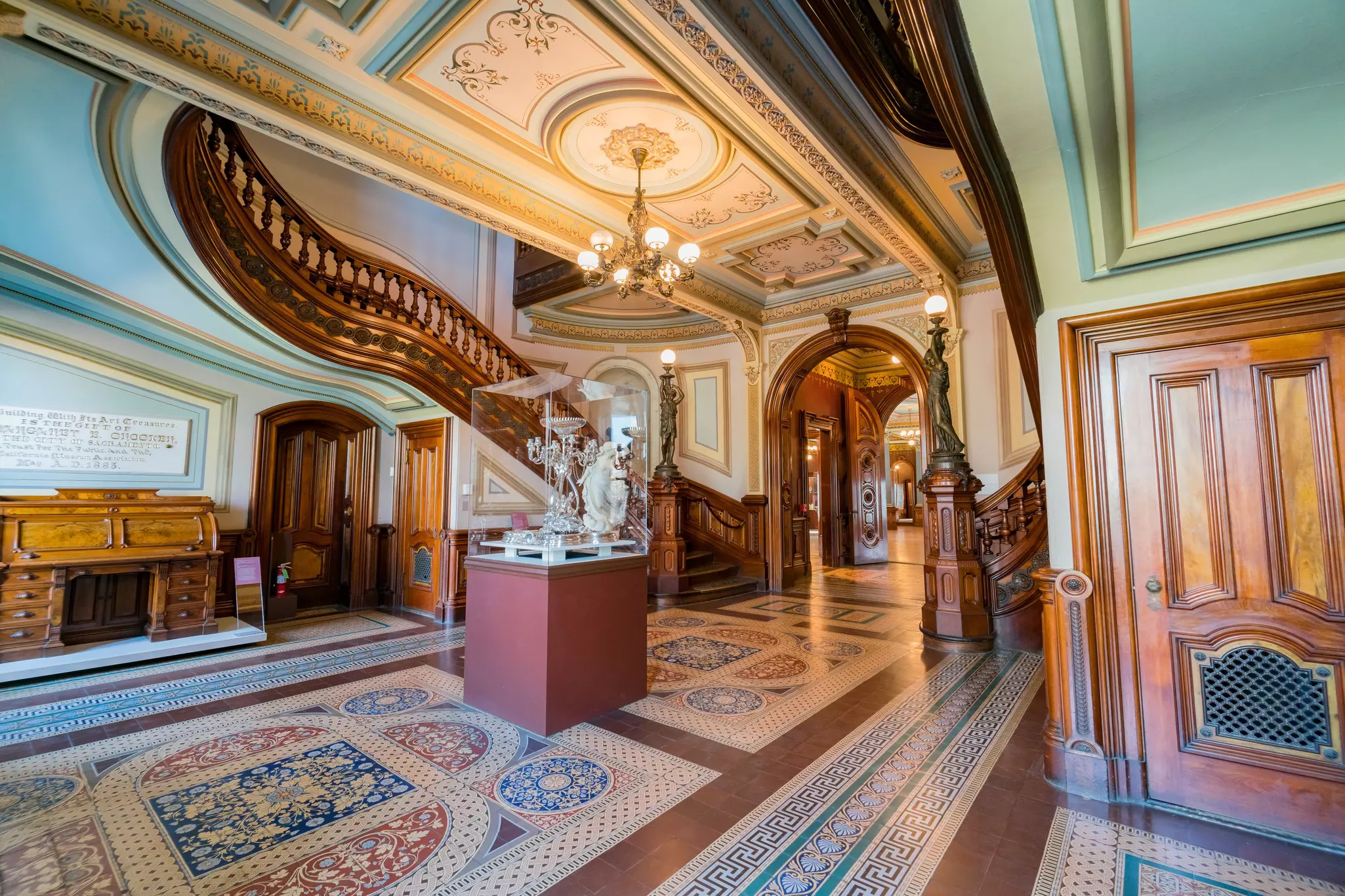An entrance hall with intricately tiled floor, a decorative ceiling with chandelier and elaborate bannister on its curved staircase.
