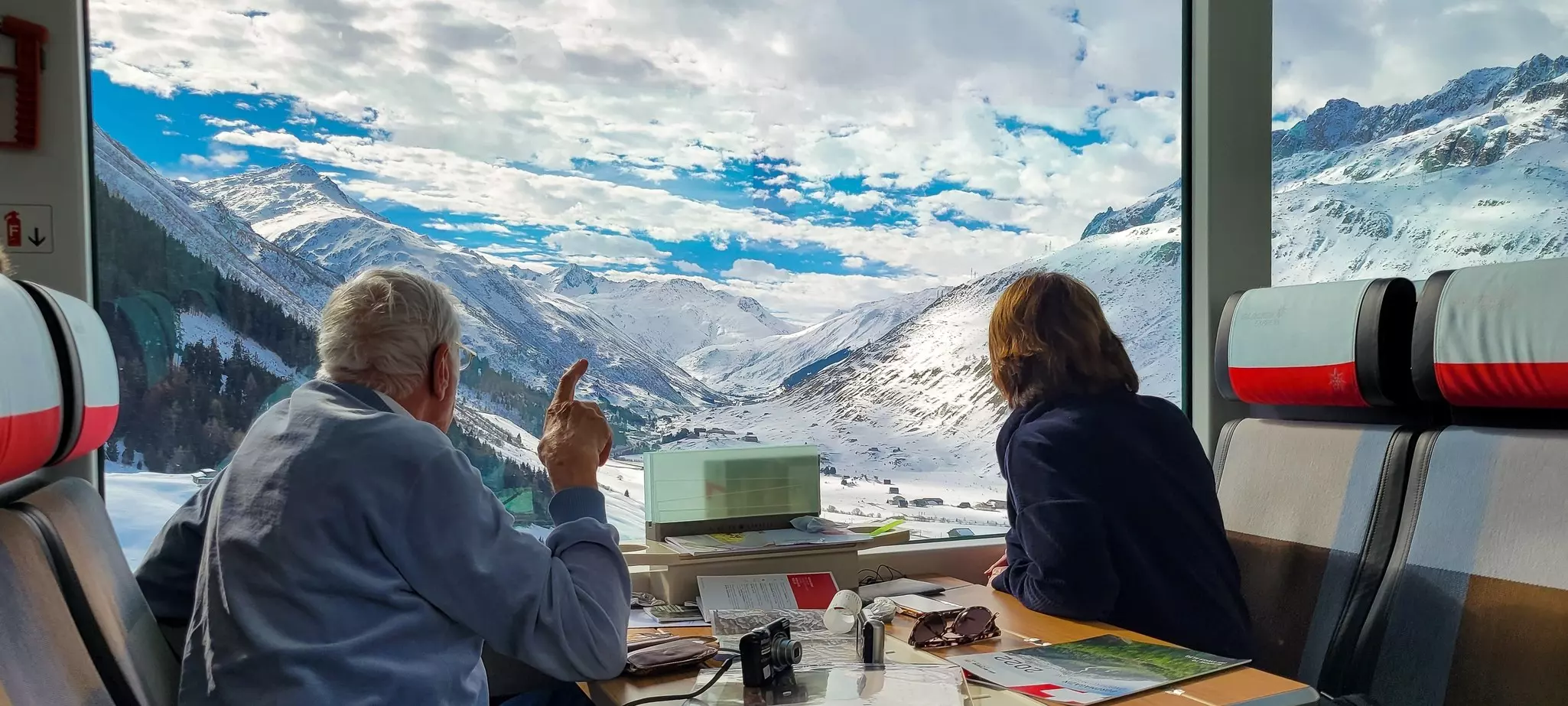 Two people sit on a train looking out a large window at snow-covered mountains in Switzerland.