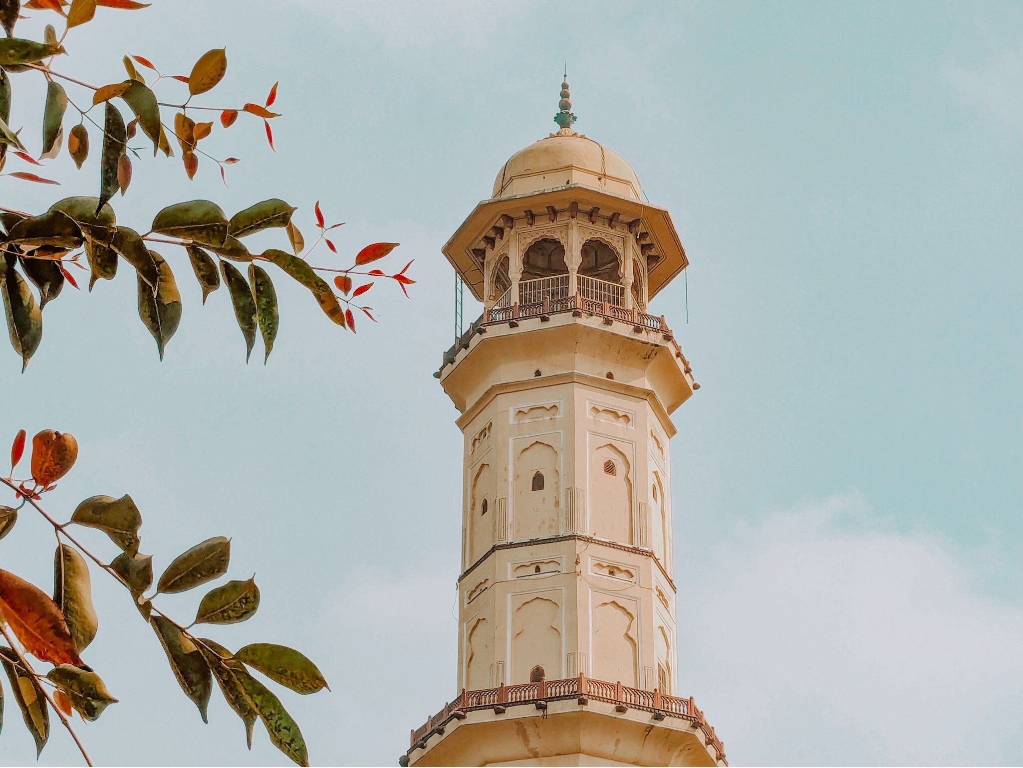 Sargasuli Tower (Isarlat) standing against a clear blue sky on a sunny morning in Jaipur, Rajasthan, India. Historic victory tower in the Pink City with Rajput and Mughal architecture, framed by tree leaves in the foreground.