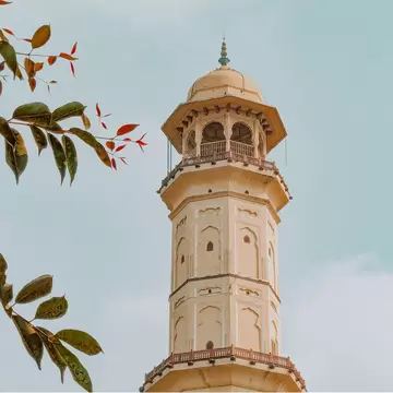 Sargasuli Tower (Isarlat) standing against a clear blue sky on a sunny morning in Jaipur, Rajasthan, India. Historic victory tower in the Pink City with Rajput and Mughal architecture, framed by tree leaves in the foreground.
