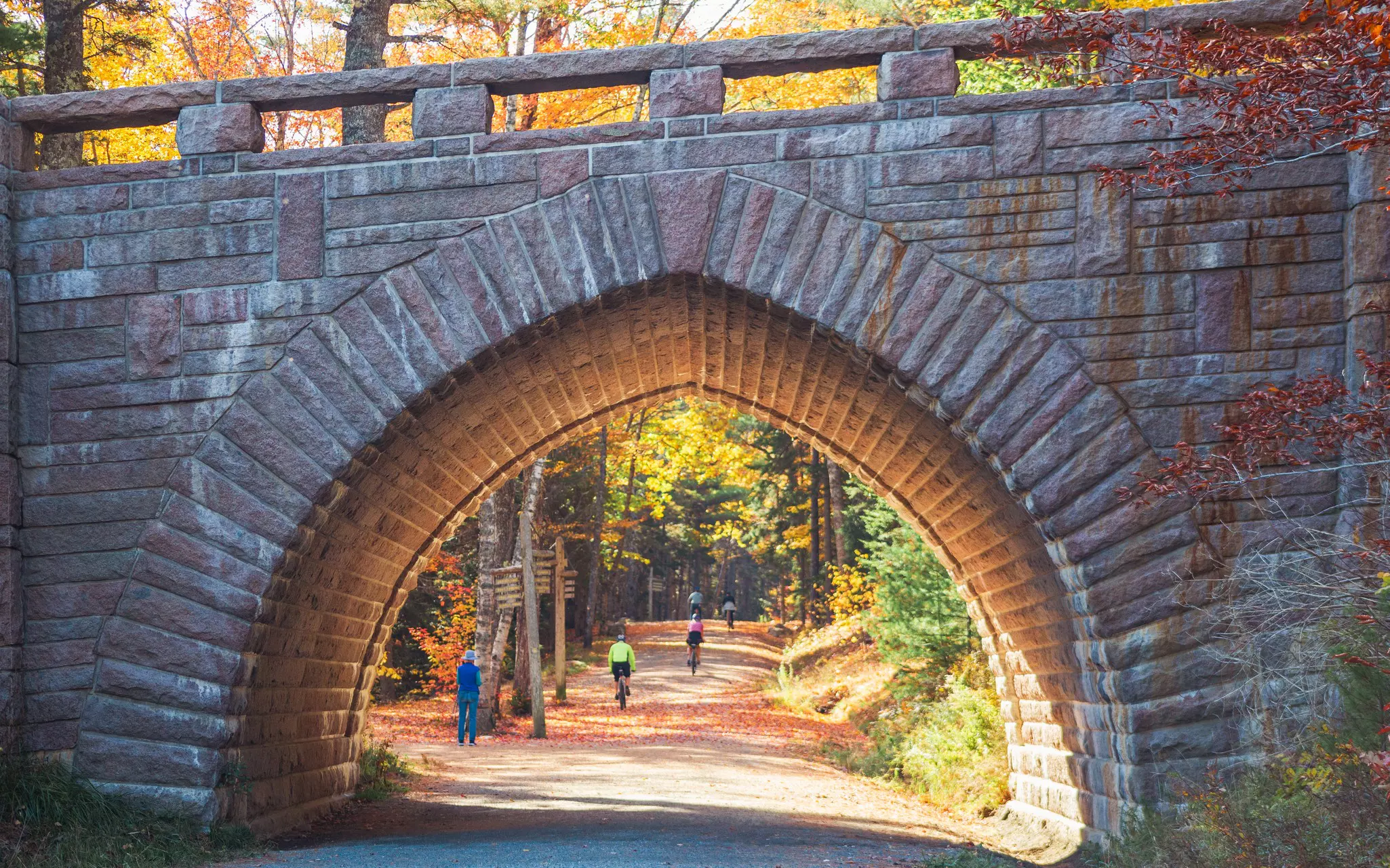A stone bridge crosses a car-free gravel road in Acadia National Park. Cyclists pedal along the wooded trail.
