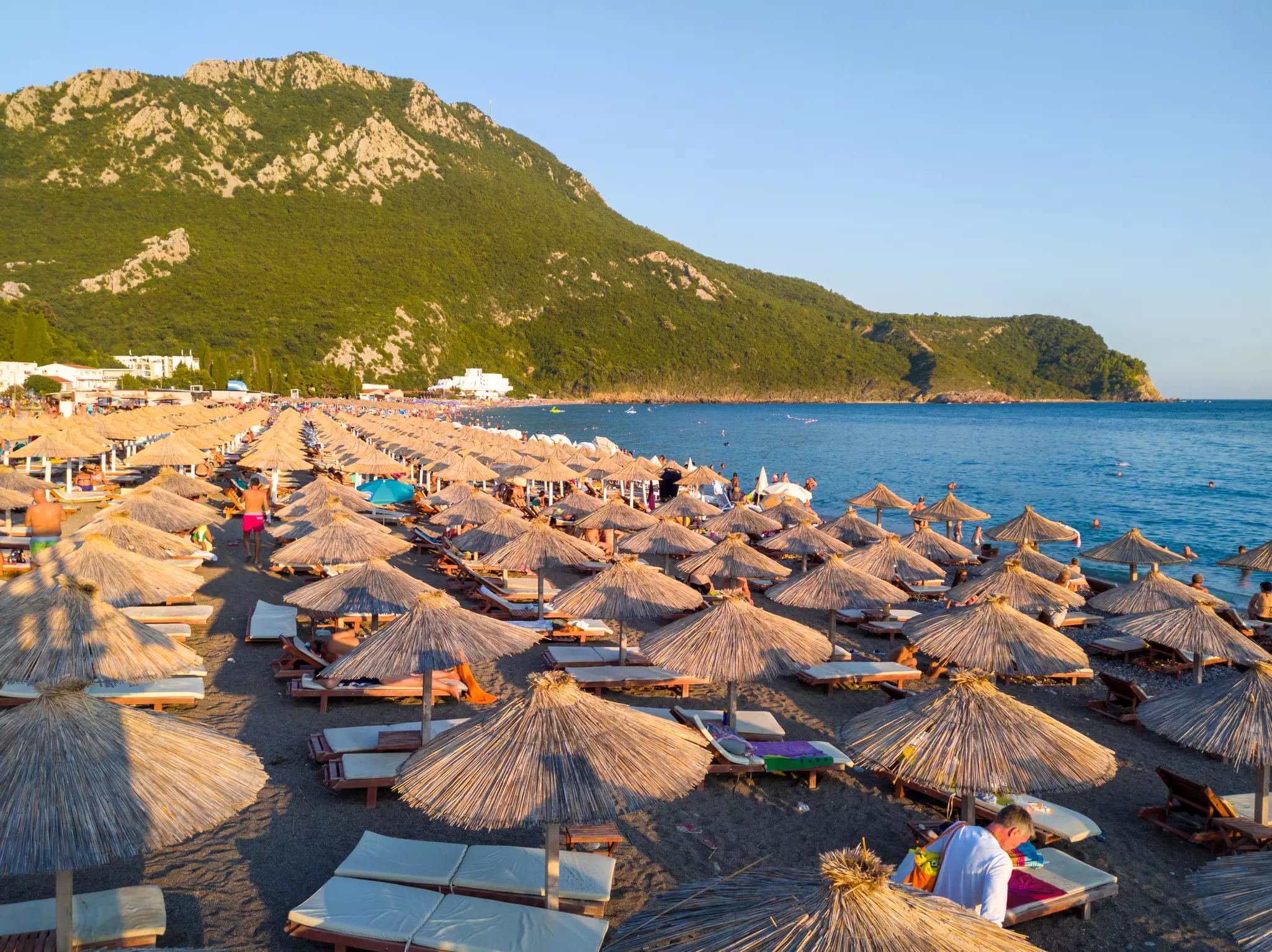 A beach is filled with dozens of thatched sun umbrellas, providing shade to people lying on loungers under them.