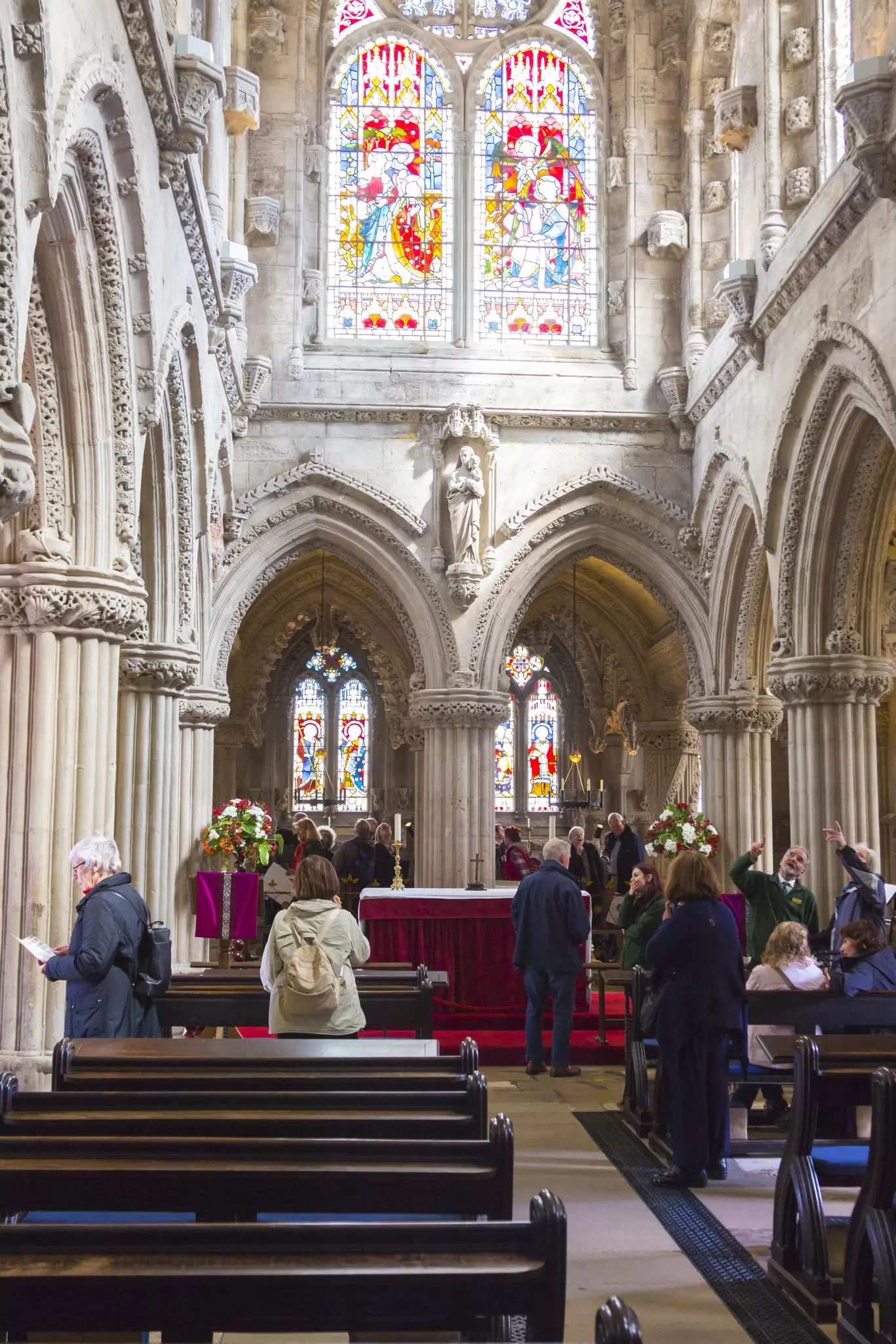 Visitors in pews at a chapel with ornate stained glass windows above them