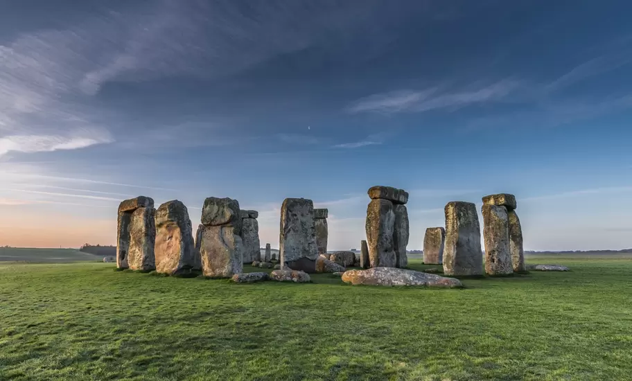 Stonehenge stone circle in Wiltshire on green grass, standing sarsens and lintels lit by low sun under a blue sky