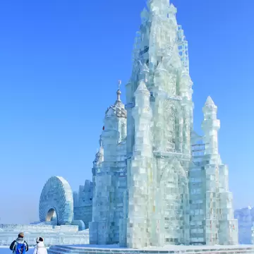 Tall spired castle built from translucent ice blocks on snowy ground under a clear blue sky, two people walking in front.