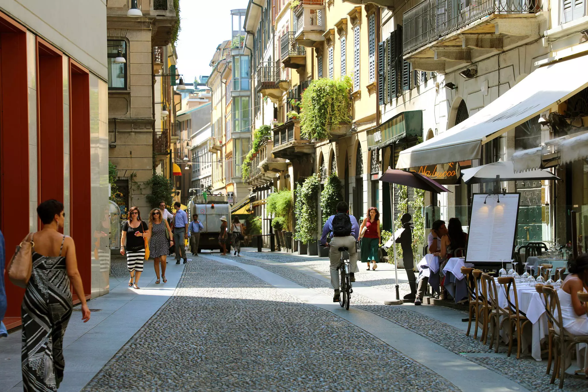 A cafe with white tablecloths, a biker and walkers on a street in Milan.