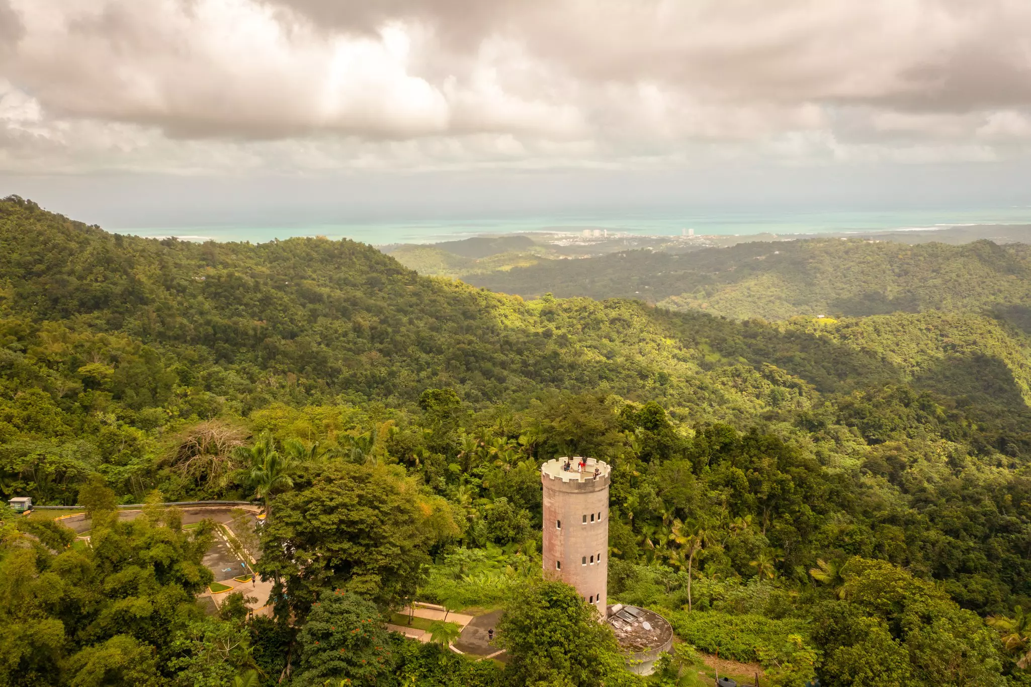 A stone tower with turrets stands in a vast green forest; turquoise water is on the horizon.