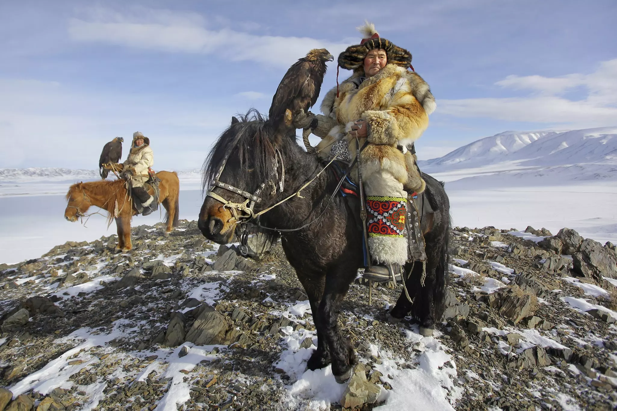 Two people in fur clothing atop horses and holding golden eagles atop a rocky mountain with snow-capped peaks in the distance.