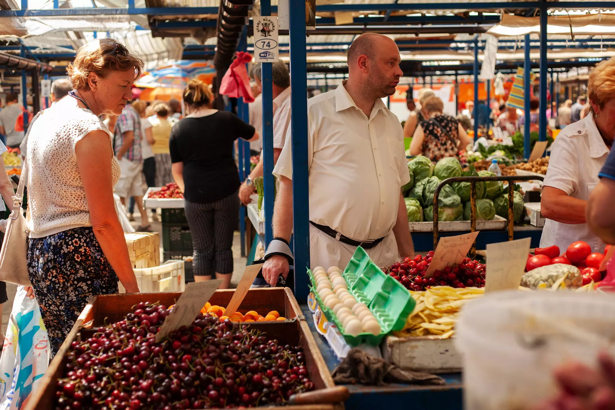 Shoppers at a produce stand in a open market; there is a large bin of cherries and open containers of eggs.
