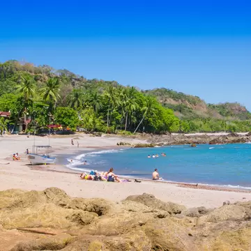 Golden sand curves past palms and a moored boat to a rocky point, with swimmers in blue water and rocks in the foreground