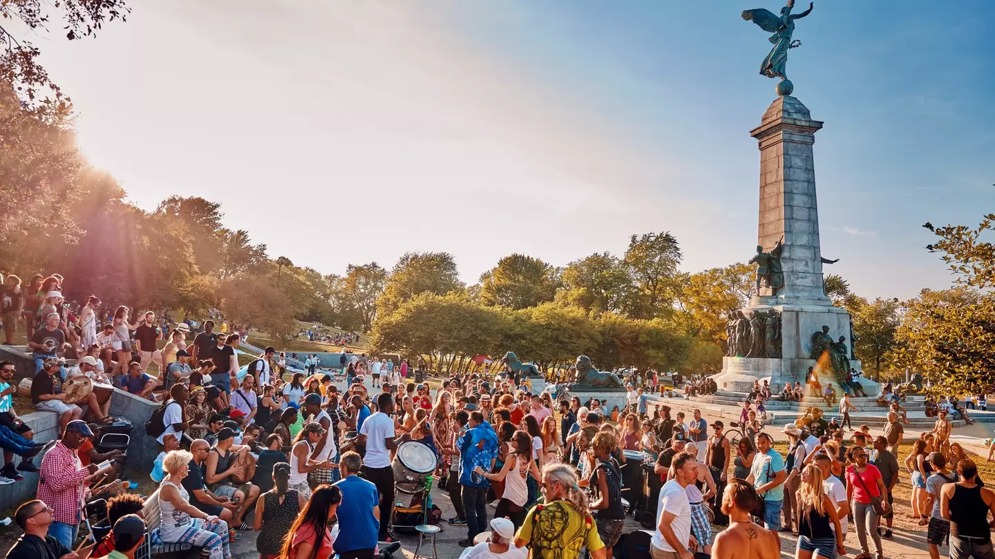 A diverse crowd of people gathered and cheering around a tall monument in a park. Some people are beating drums