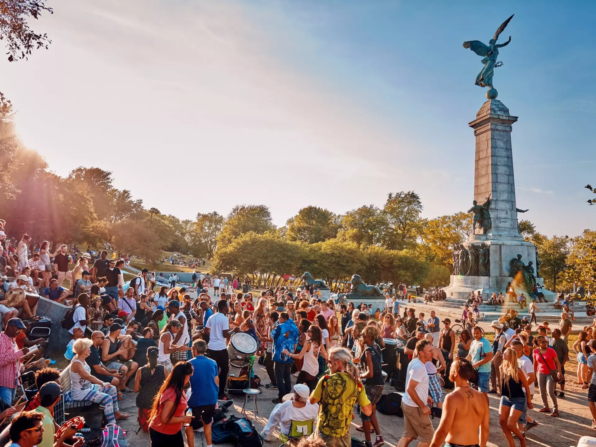 A diverse crowd of people gathered and cheering around a tall monument in a park. Some people are beating drums