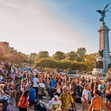 A diverse crowd of people gathered and cheering around a tall monument in a park. Some people are beating drums