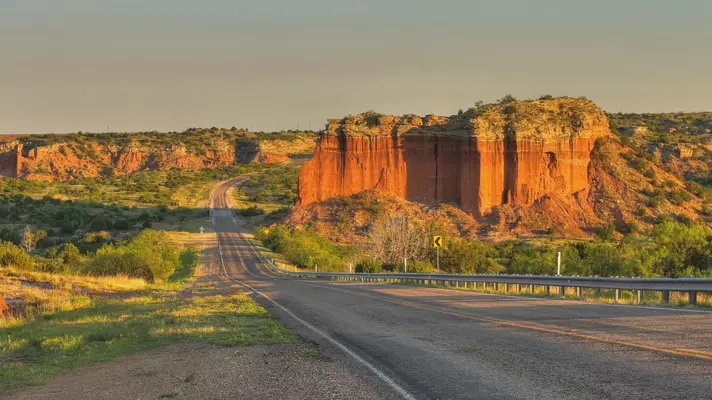 A road leading through large red rock formations is seen at sunset.