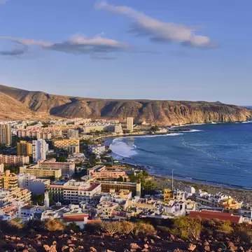 Los Cristianos, Tenerife, from a rocky hilltop: pastel apartment blocks beside a dark-sand beach and a brown headland.