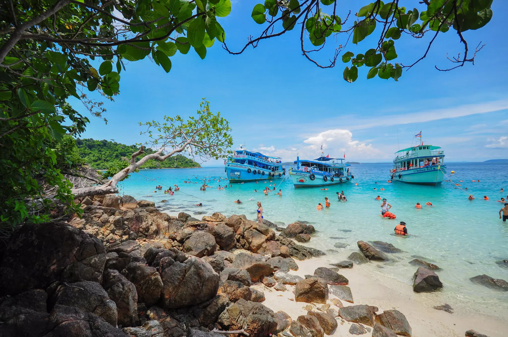 People, some wearing lifejackets, swim and snorkel off the coast of a tropical beach. Three boats are anchored in the water just offshore.