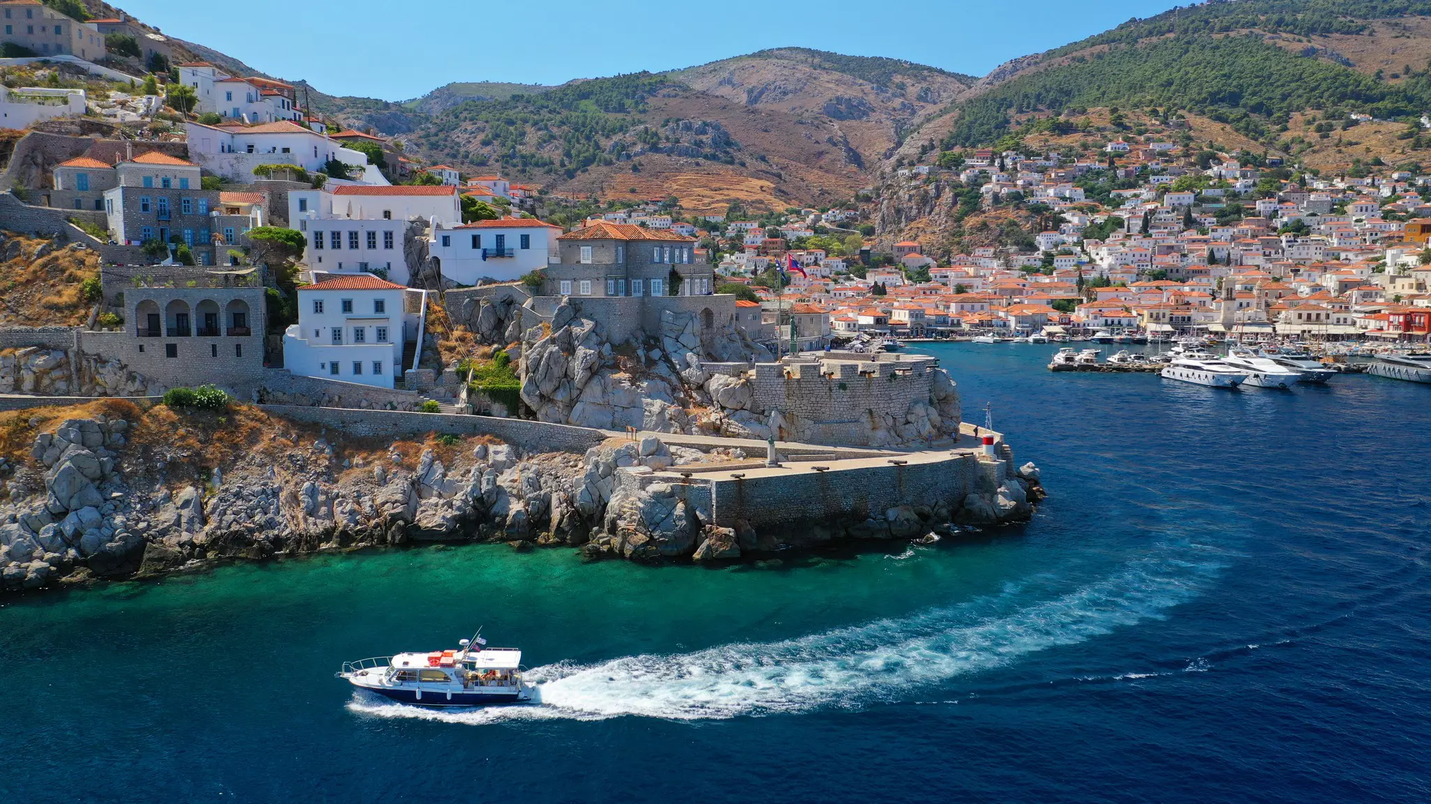 A wide view of a boat leaving a harbor, trailing a white wake as it cruises in the water along the coast.