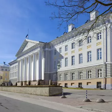 White neoclassical facade with a tall columned portico and gold window ornaments under a clear blue sky
