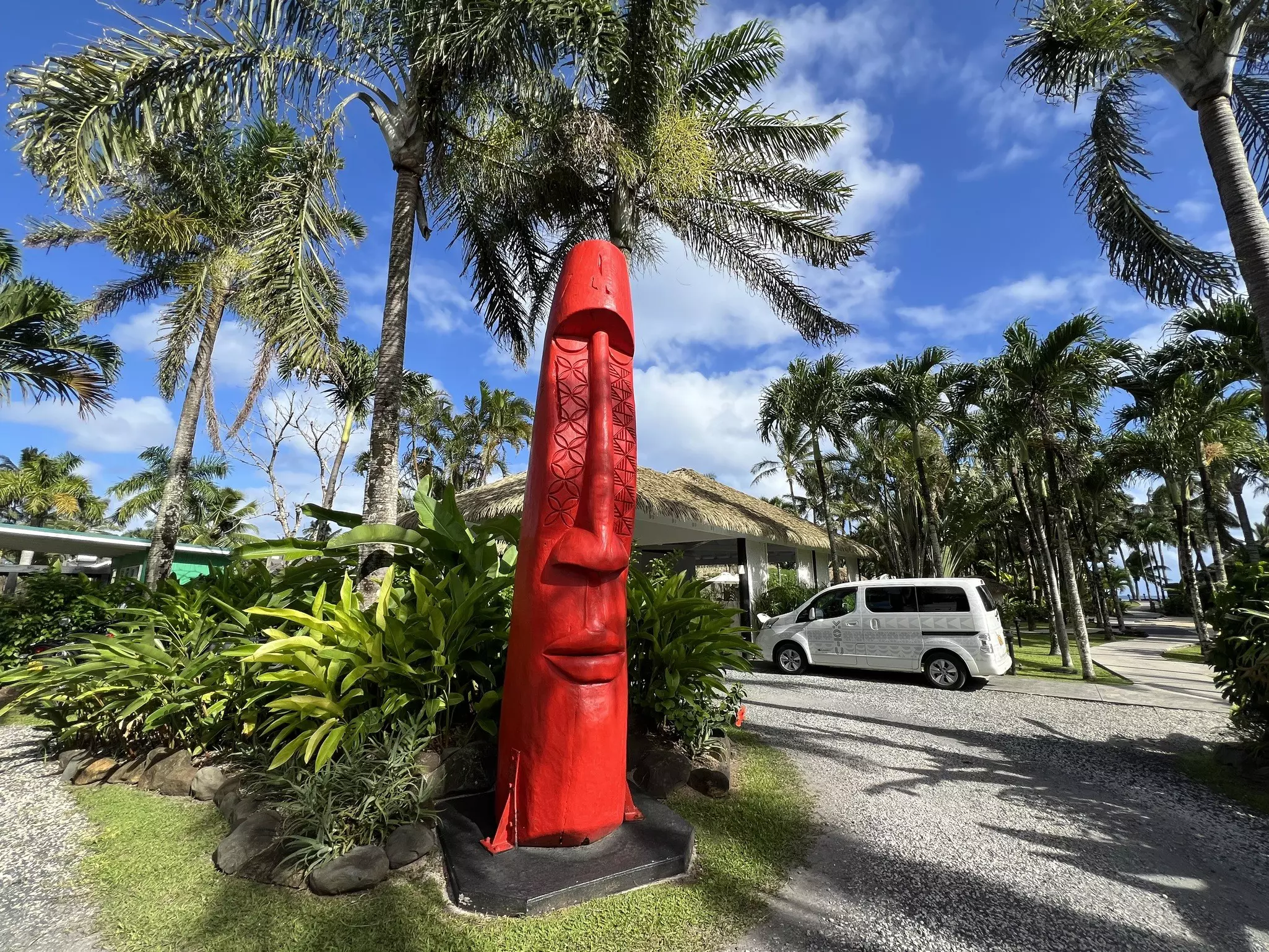 A large red sculture of an elongated head and face stands outside the entrance to a hotel surrounded by palm trees.