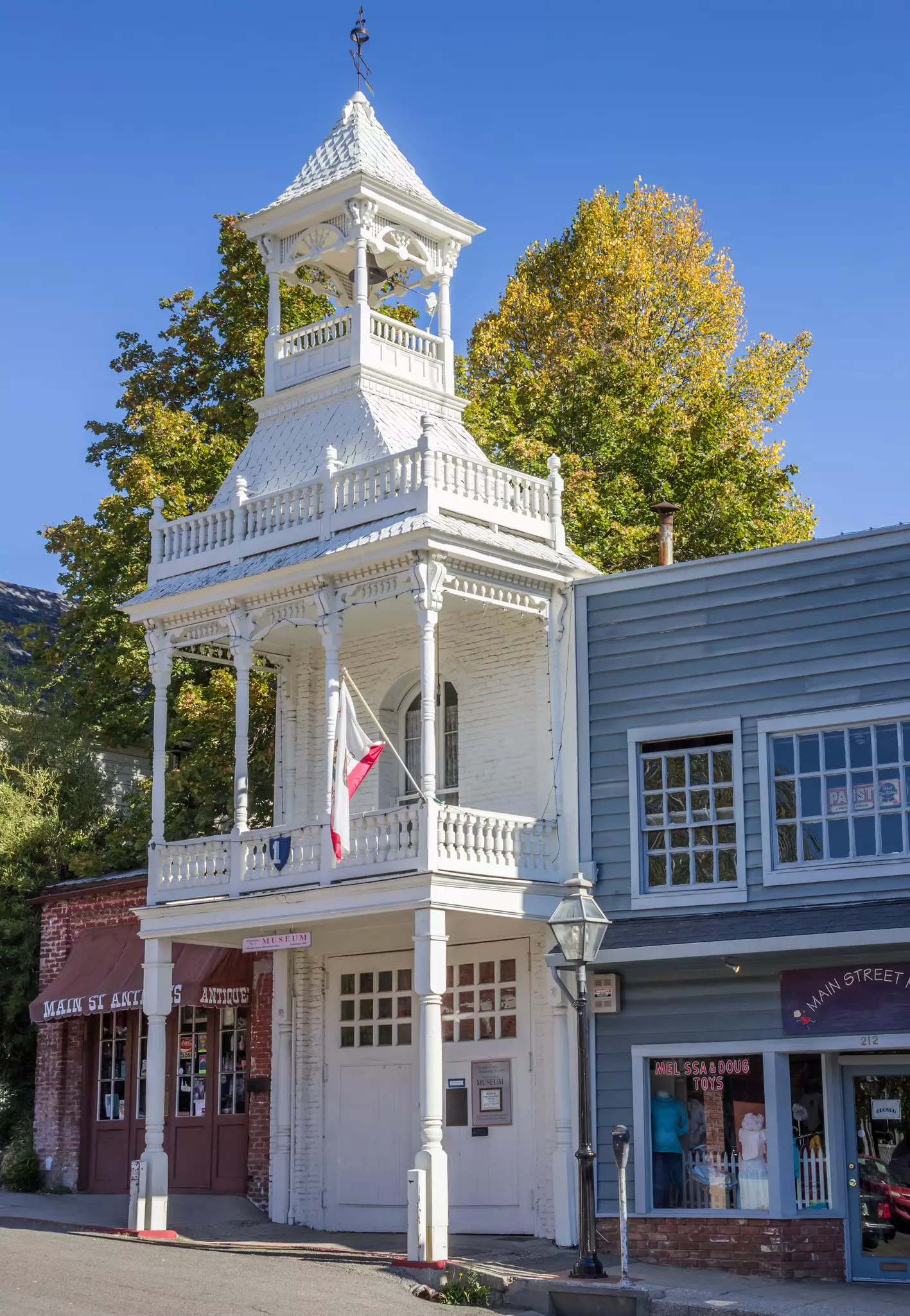 A historic wooden firehouse with quaint stores either side in a Victorian-era Main St.