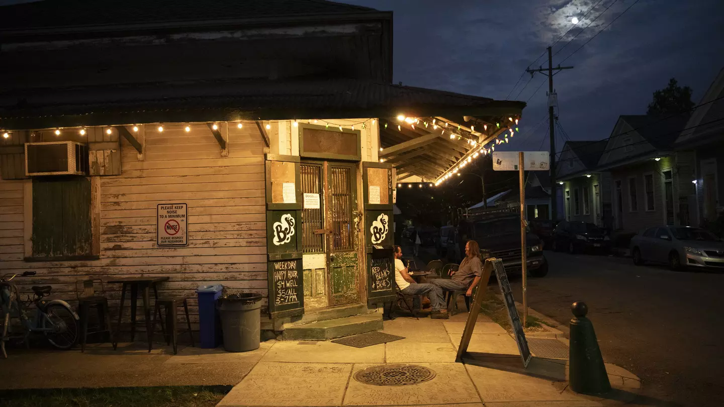 People sit outside at dusk under the awning of a bar in a city.