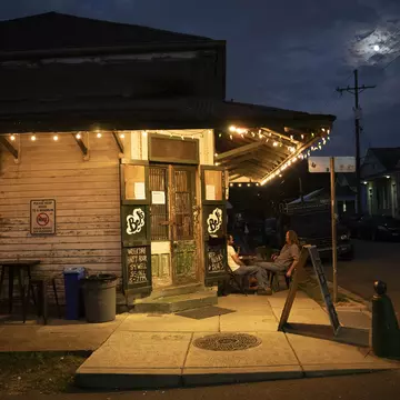 People sit outside at dusk under the awning of a bar in a city.
