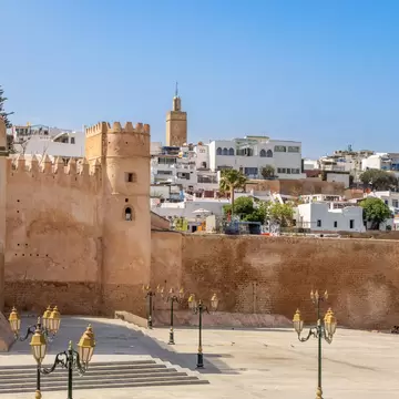 Lamps on a plaza by ancient stone walls and towers; a city with white buildings is above on a hillside.