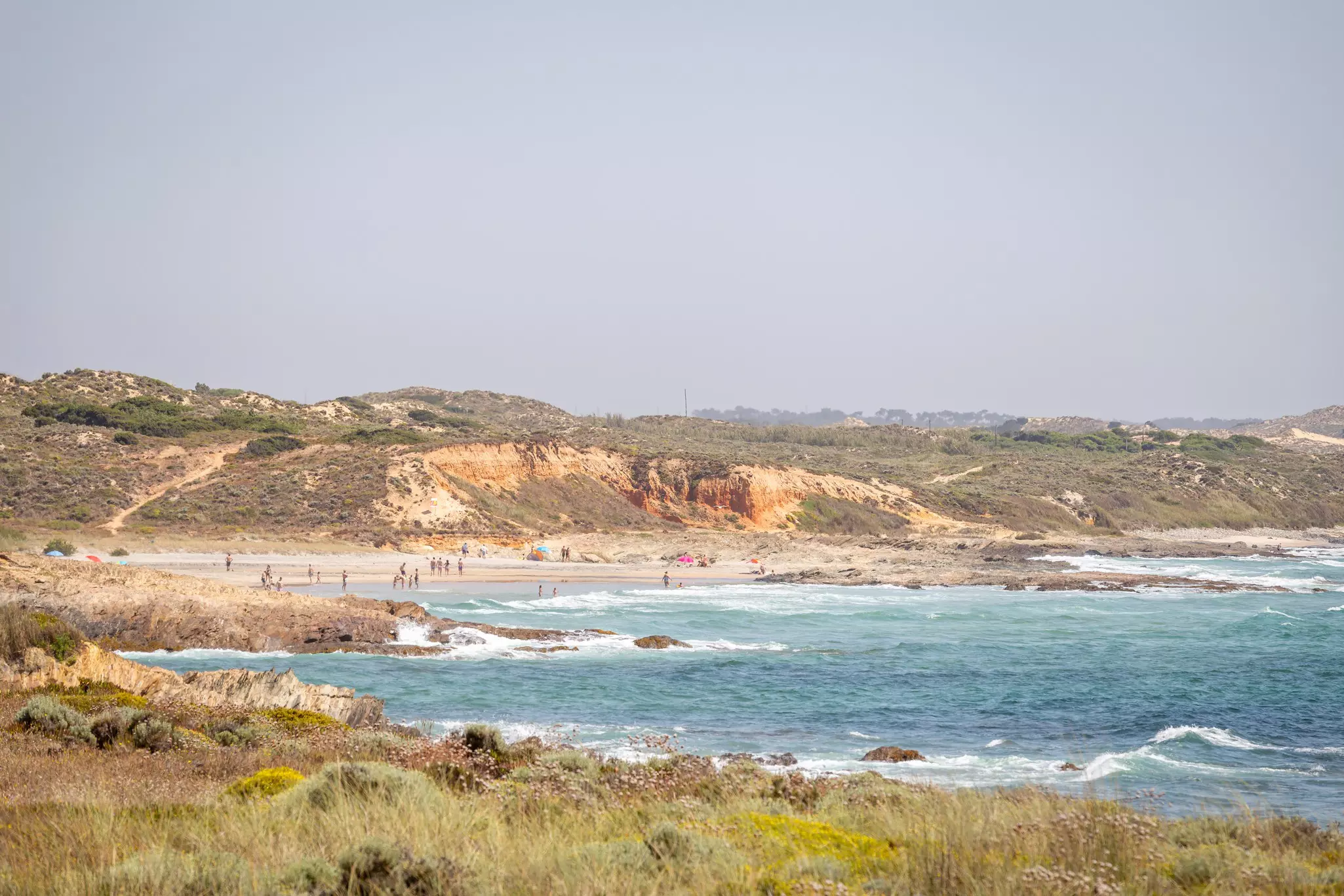 A series of rolling sandy cliffs give way to a beach where people are playing in the surf.