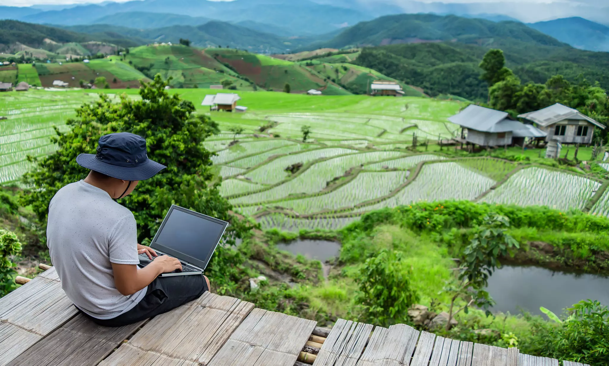 A man wearing a bucket hat uses a laptop with rice fields and mountains in the background.