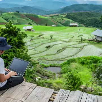 A man in a sun hat works on a laptop on a wooden deck overlooking green rice terraces and hills.