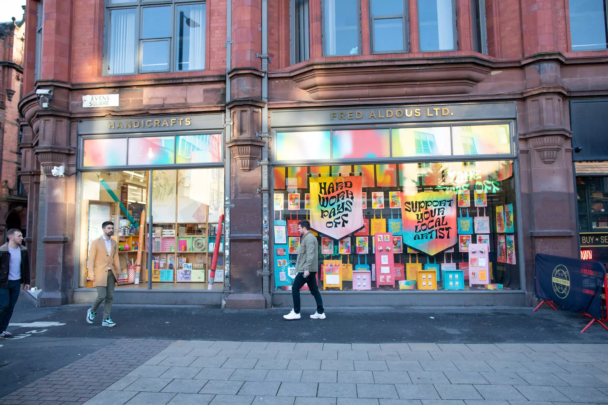 People pass a crafts store with a colorful window display and a sign saying "support your local artist"