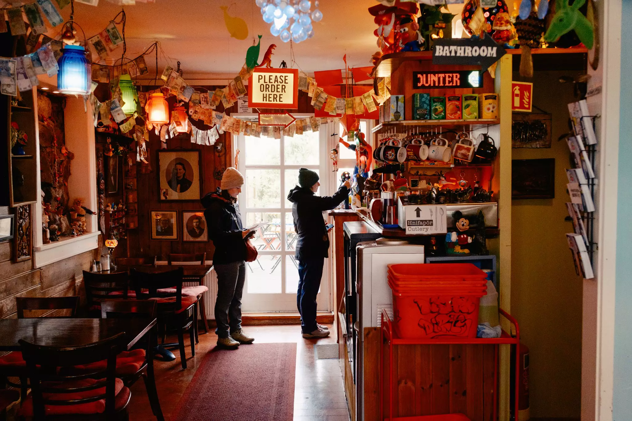 People at a counter by a door in a small cafe with tables to one side.