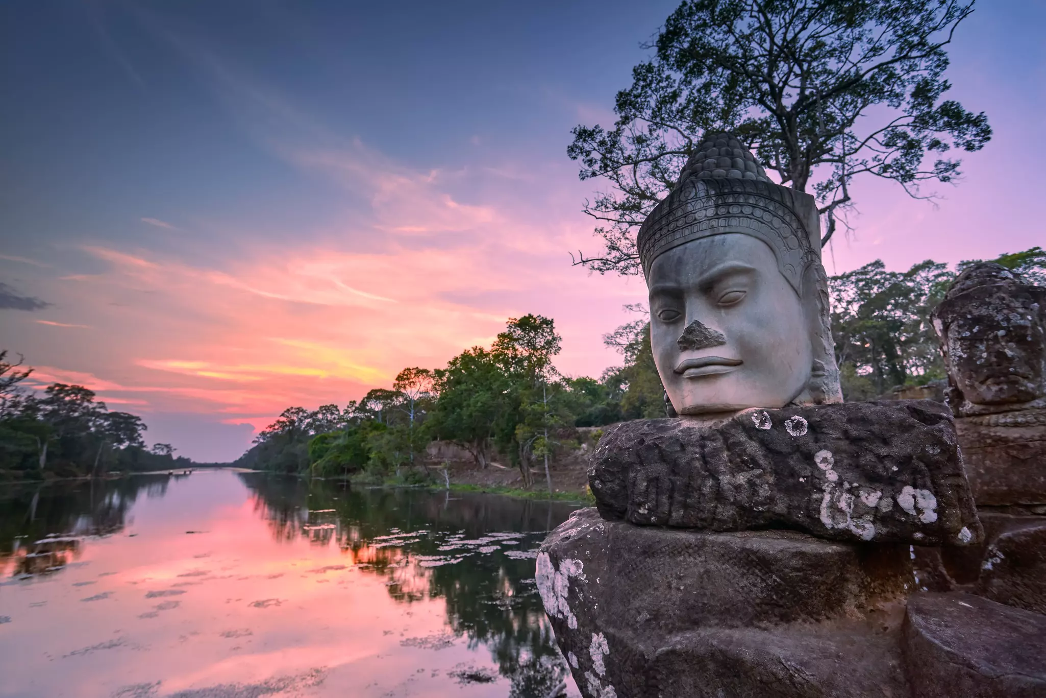 A giant sculpture of a head with a serene expression sits on a rock by water near Siem Reap, Cambodia, at sunset.