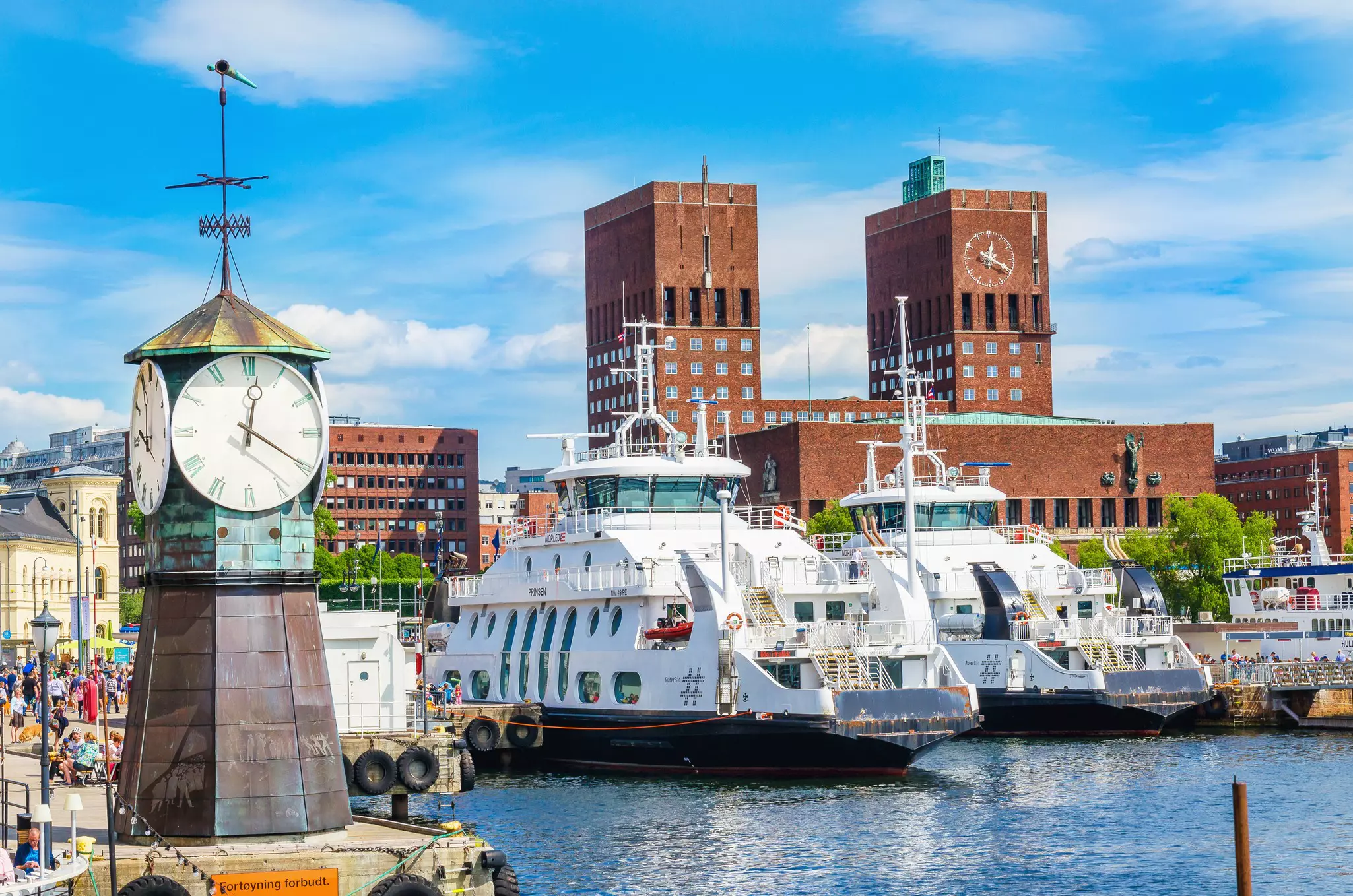 Large clock structure on a cement dock along a harbor with ferries docked to the right and a large brick two-towered building in the distance on a sunny day.