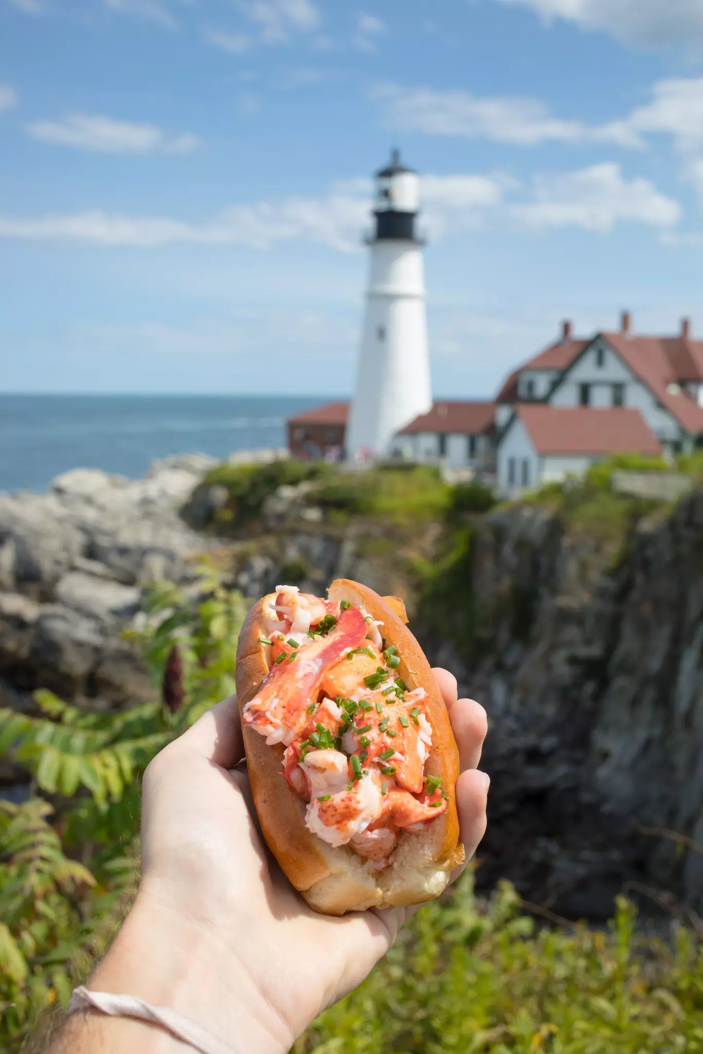 A hand holds out a bread roll stuffed with lobster in a coastal setting near a lighthouse.