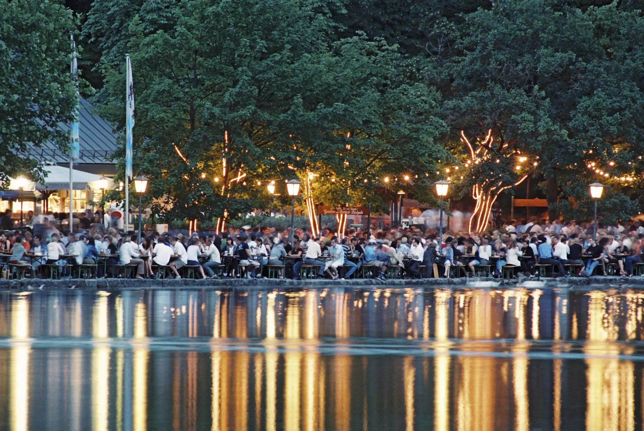 People are seen sitting at tables at an outdoor beergarden in the evening. Lights are reflected in a lake in the foreground.