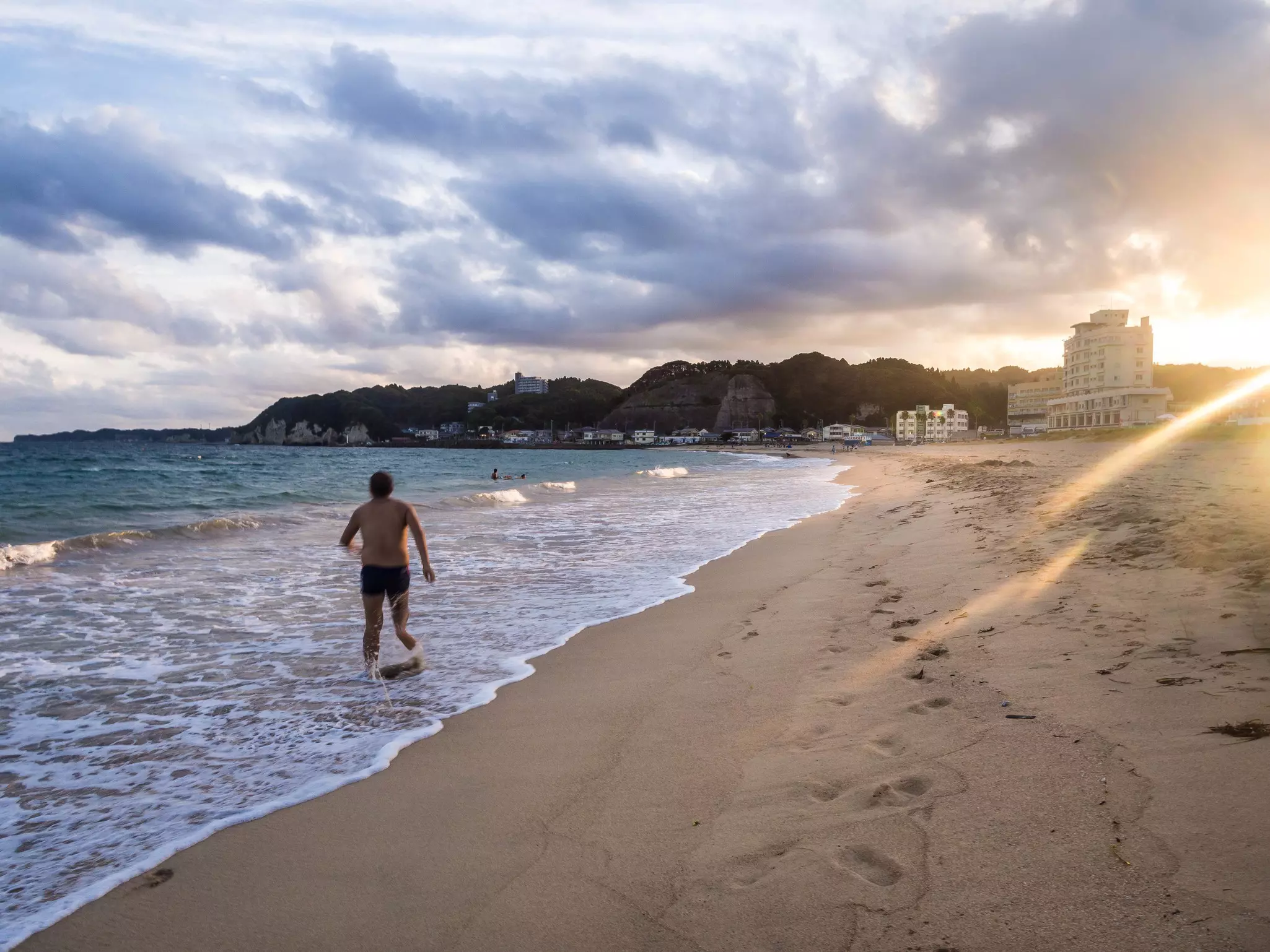 A man wades into the surf on a beach at sunset.