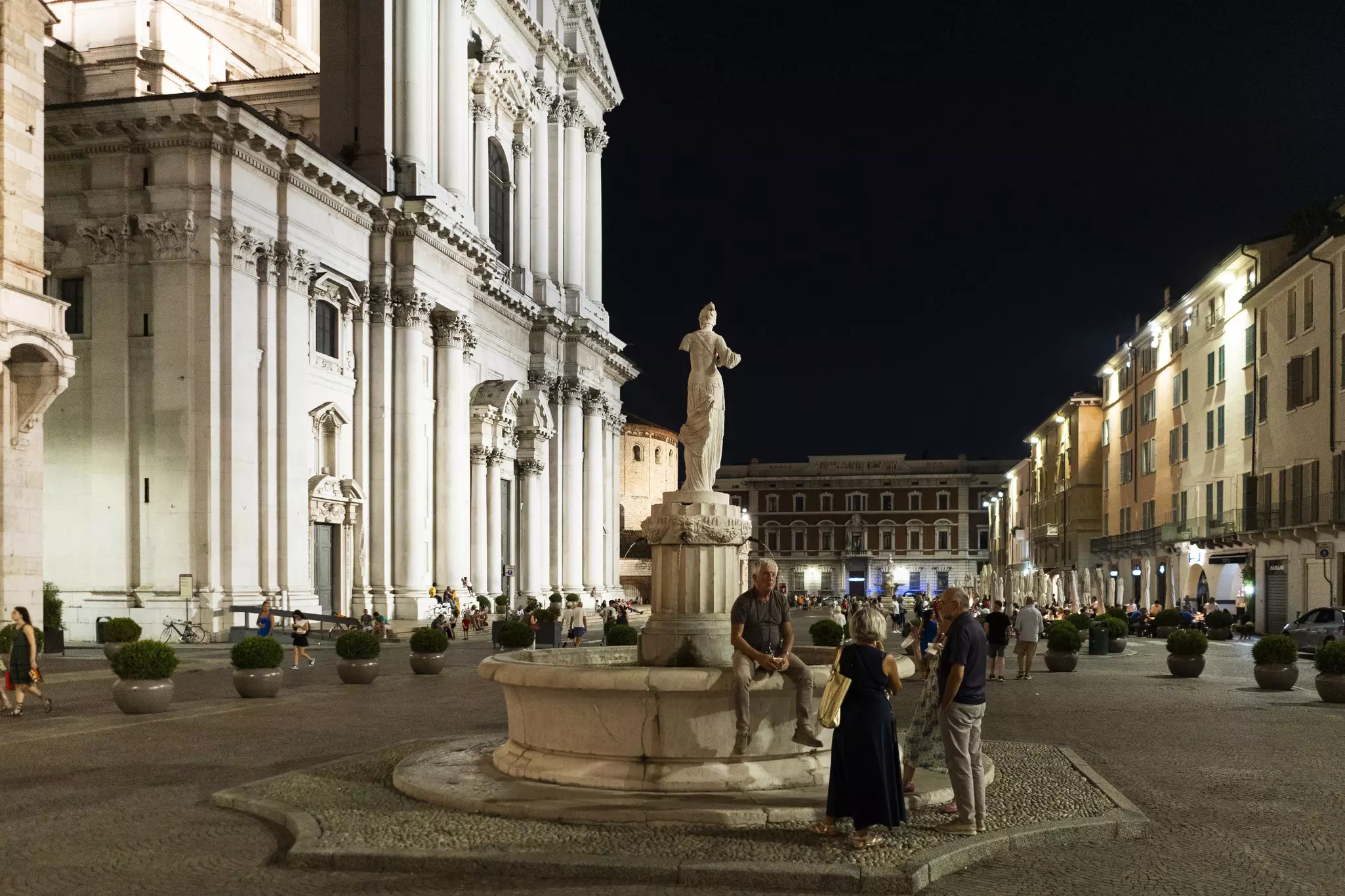 A group of people sit around a fountain having wine in a large Italian piazza.