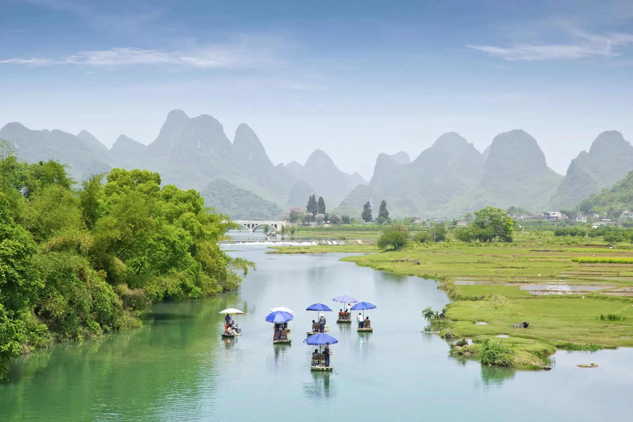 People in boats with umbrellas are on a waterway that leads to peaks in silhouette in the distance.