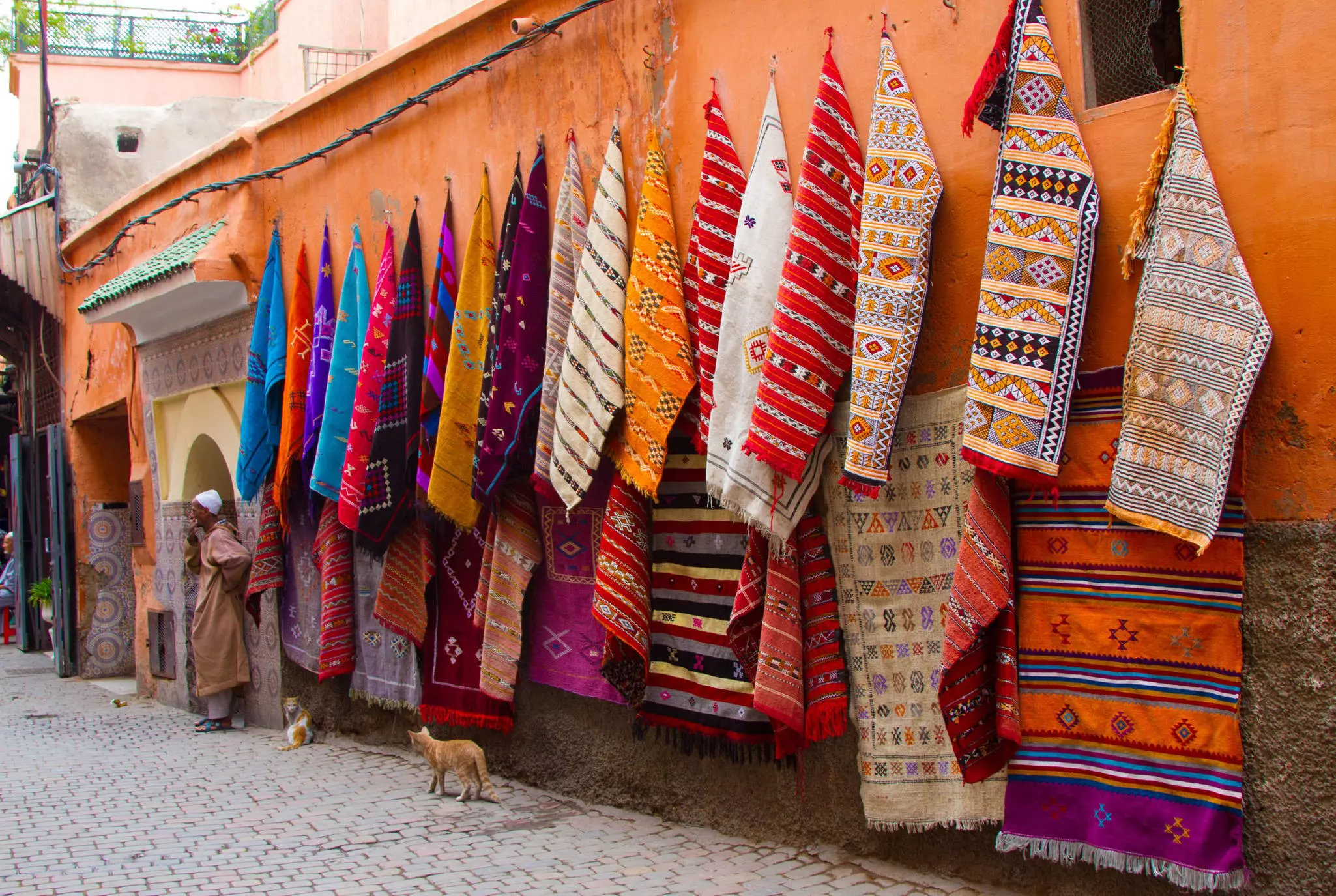 Colorful rugs hanging on a brightly painted orange wall in an alleyway within a marketplace.