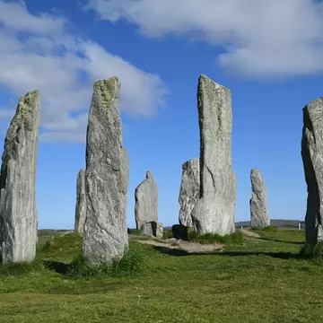 Tall grey lichen-covered standing stones in a ring on green grass at Callanish, Isle of Lewis, under a blue sky with clouds.