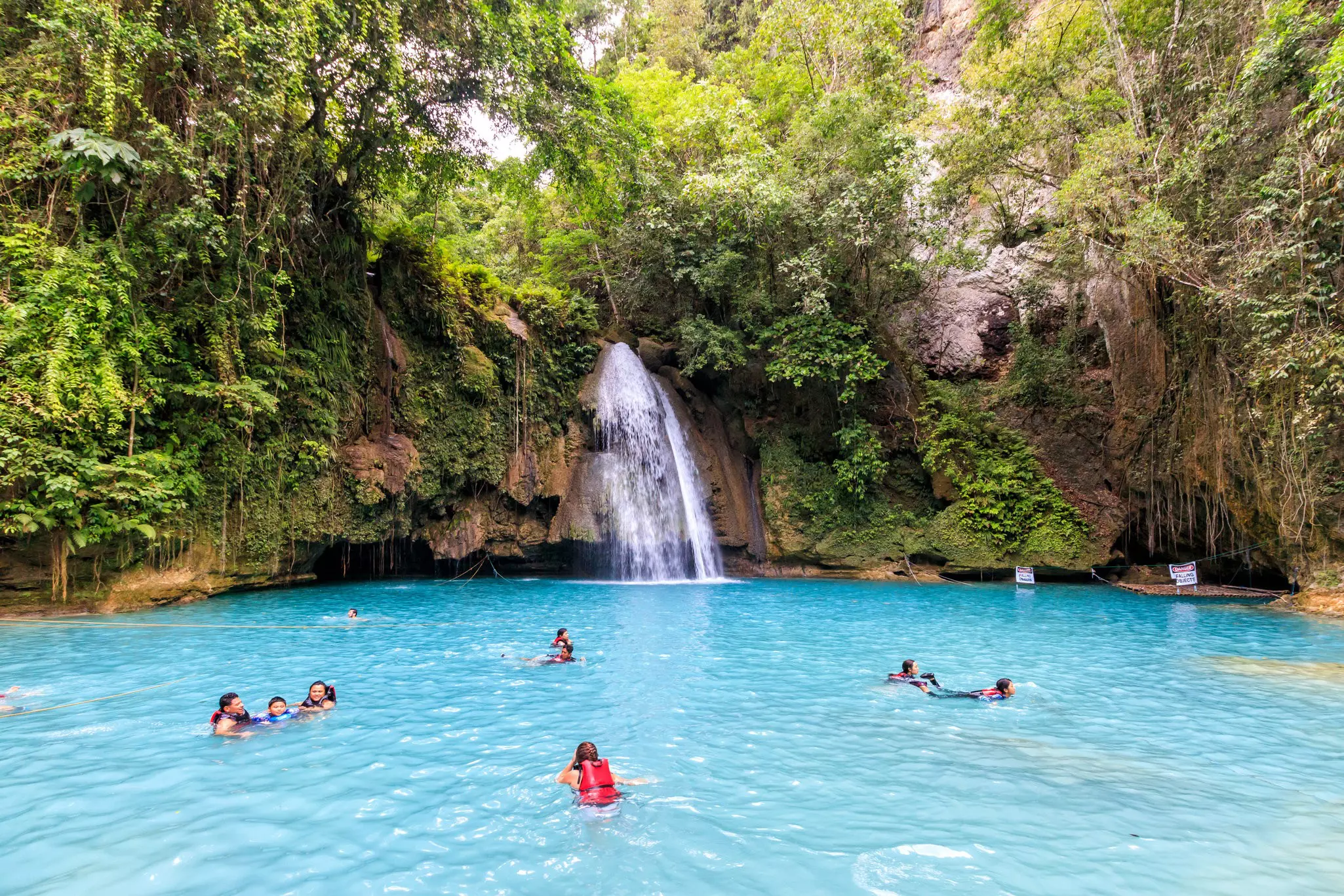 A shot of the blue pool at the bottom of Kawasan Falls, Cebu with a few people swimming in it. The blue waters are surrounded by lush greenery and we can also see the small waterfall.