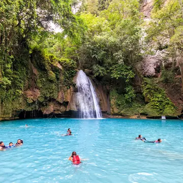 A shot of the blue pool at the bottom of Kawasan Falls, Cebu with a few people swimming in it. The blue waters are surrounded by lush greenery and we can also see the small waterfall.