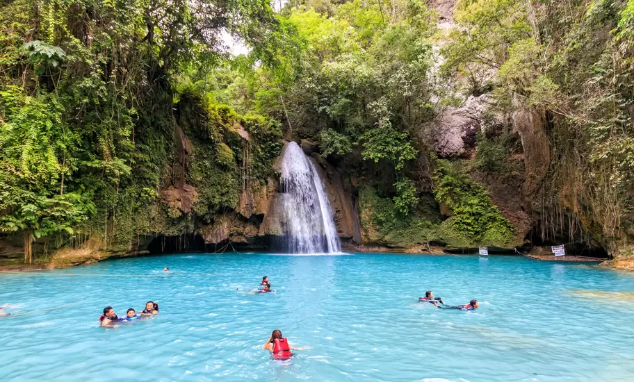 A shot of the blue pool at the bottom of Kawasan Falls, Cebu with a few people swimming in it. The blue waters are surrounded by lush greenery and we can also see the small waterfall.